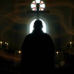 Bild eines Priesters, der mit dem Rücken zum Kreuz in einer Kirche in einem dunklen Raum betet