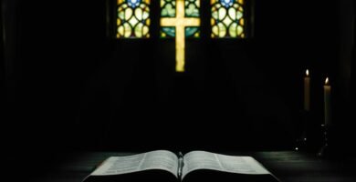Bild einer offenen Bibel in einem von einem Kreuz beleuchteten Raum neben Kerzen in einem dunklen Raum