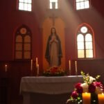 Bild des Inneren einer Kapelle mit dem Bild der Jungfrau Maria um Kerzen und Blumen herum, durch die Fenster fällt Licht